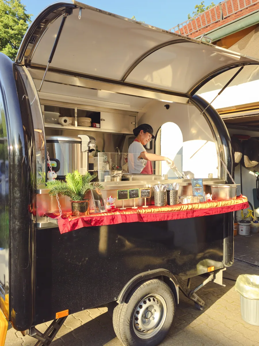 Der kleine schwarze Foodtrailer mit hochgeklappter Klappe, Paew kocht darin am Herd