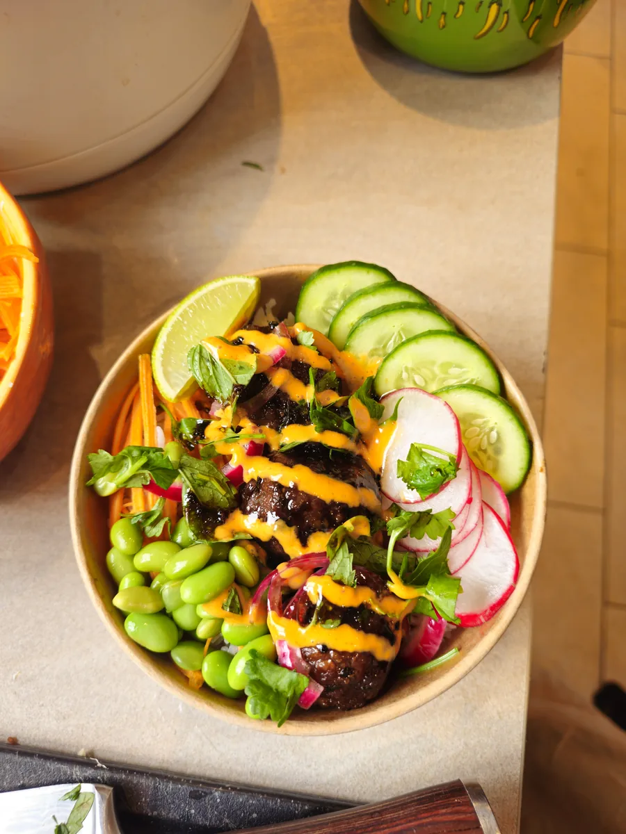 Veggie Bowl Hanoi in einer Pappschale: Reis mit veganen 5-Spice-Hackbällchen in Hoisin-Glasur, Erdnuss-Sesam-Dip mit Gochugaru, Edamame, Gurke, Radieschen, Karotte, frischen Kräutern und Limette